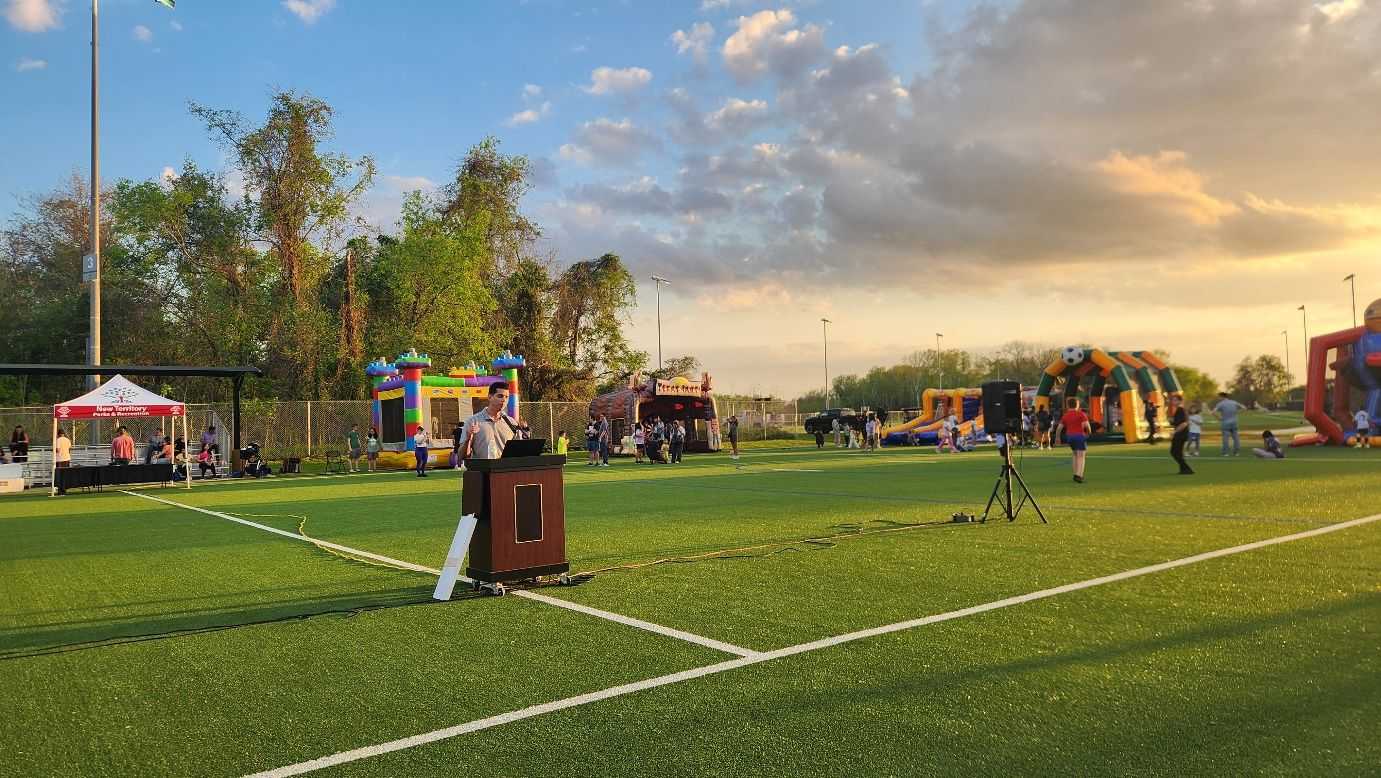 Outdoor event on a turf field with a podium, speaker, inflatable play structures, and people gathered under a colorful sunset sky.