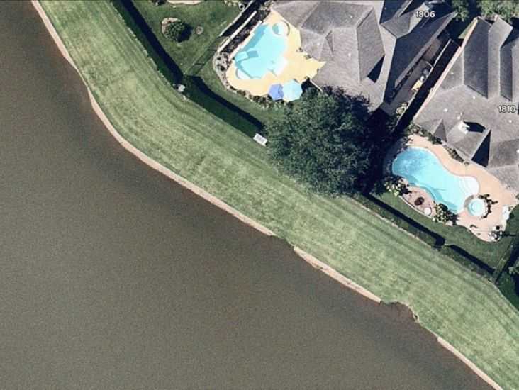 Aerial view of lakeside homes with backyard pools and a sloped grassy embankment separating the properties from the water.